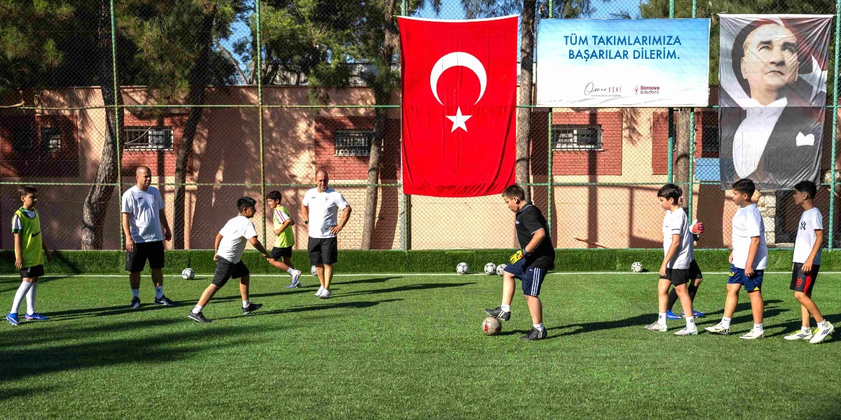 Açık havada, suni çim kaplı bir futbol sahasında antrenman yapan bir grup çocuk ve iki antrenör. Çocuklardan biri topu sürerken, diğerleri onu izliyor. Sahayı çevreleyen tel örgülerin üzerinde büyük bir Türk bayrağı, 