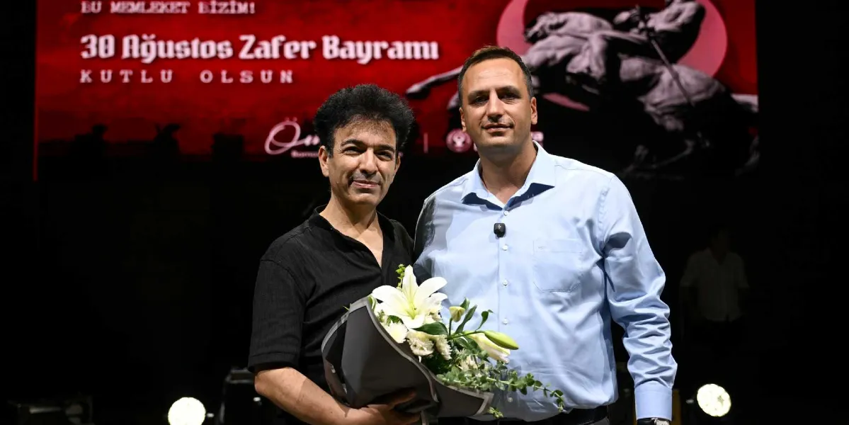 Siyah tişörtlü bir erkek, elinde beyaz zambaklardan oluşan bir buketle, açık mavi gömlekli başka bir erkeğin yanında durarak objektife gülümsüyor. Arka planda, üzerinde 
