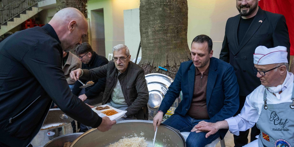 Bornova Belediye Başkanı Ömer Eşki, İzmir Makedonya Göçmenleri Kültürünü Yaşatma ve Dayanışma Derneği'nin iftar etkinliğinde, büyük kazanların başında vatandaşlara yemek servisi yapıyor. Başkan Eşki, elindeki tabakla bir vatandaşa yemek ikram ederken, yanında lacivert ceketli bir kişi ve üzerinde 