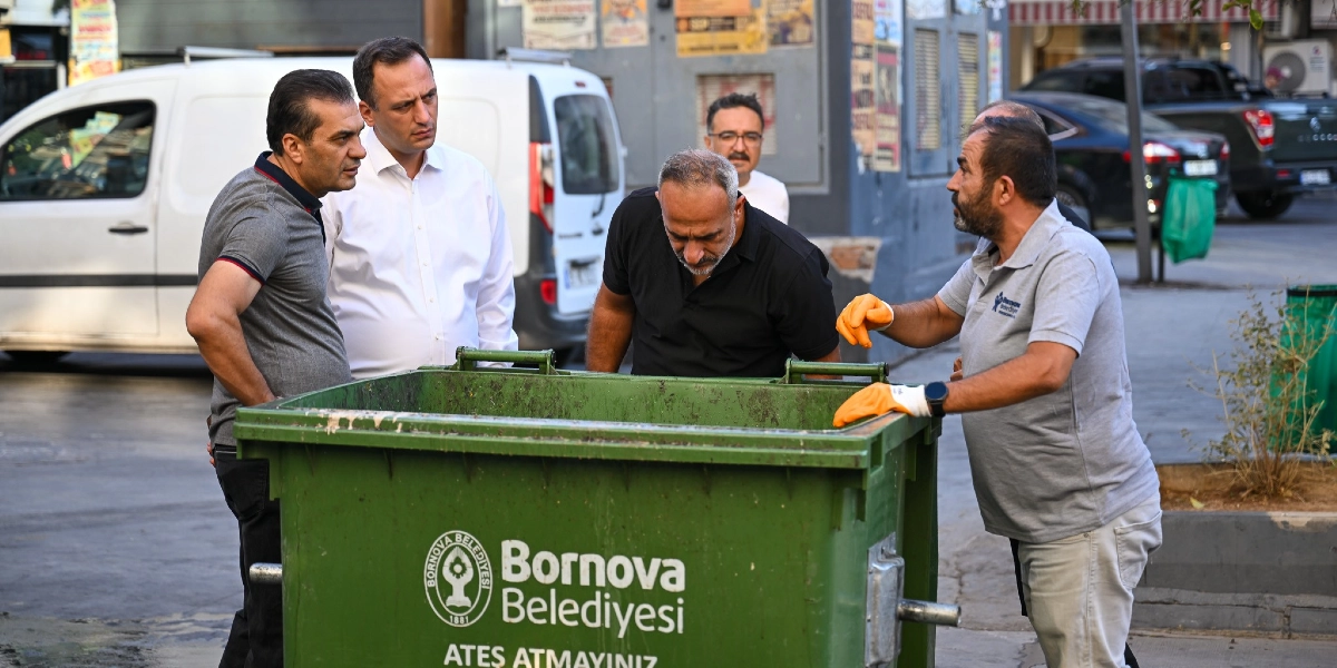 Bornova Belediye Başkanı Ömer Eşki ve beraberindeki heyet, üzerinde Bornova Belediyesi logosu bulunan yeşil bir çöp konteynerini inceliyor. Başkan Eşki siyah tişörtüyle konteynerin içine bakarken, yanında bulunan belediye görevlisi turuncu eldivenleriyle konteyner hakkında bilgi veriyor. Çevredeki diğer yetkililer de süreci takip ediyor.