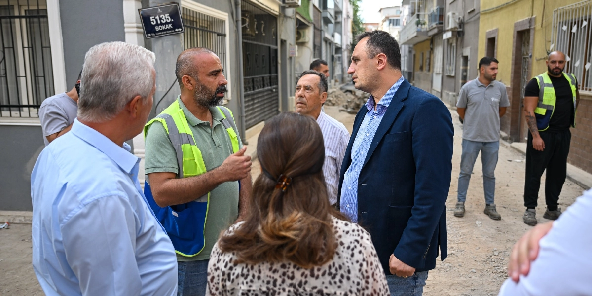 Bornova'da 5135. Sokak tabelasının bulunduğu bir inşaat alanında, üzerinde neon sarı yelek bulunan bir yetkili, lacivert ceketli bir yönetici ve beraberindeki heyetle yol çalışmaları hakkında konuşuyor. Çevredeki diğer görevliler ve vatandaşlar da çalışmaları takip ediyor.