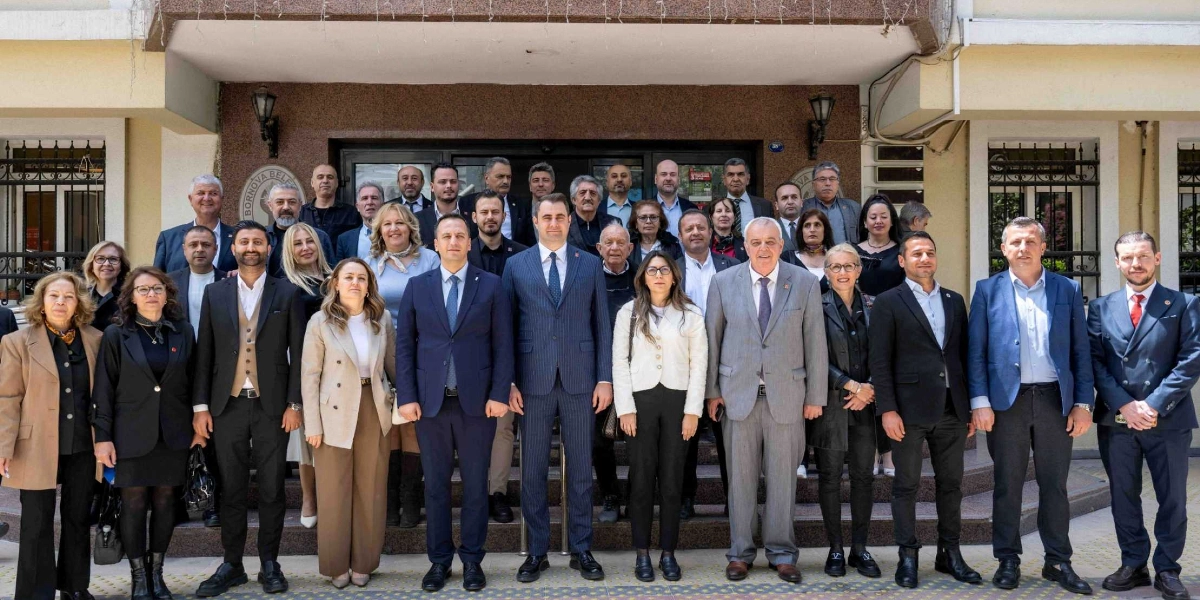 Bornova Belediyesi binasının önünde, resmi kıyafetler giymiş kalabalık bir grup kadın ve erkek siyasetçi, toplu halde poz veriyor. Ön sırada, takım elbiseli erkekler ve şık kıyafetli kadınların yer aldığı grup, merdivenlerin önünde yan yana dizilmiş şekilde duruyor. Arka planda ise binanın giriş kapısı ve pencereleri görülüyor.