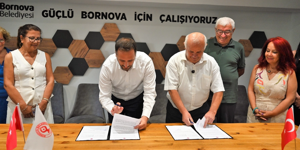 Bornova Belediyesi'nde düzenlenen bir iş birliği protokolü imza töreninde, masanın iki yanında oturan iki erkek yetkili önlerindeki belgeleri imzalıyor. Masanın etrafında ayakta duran dört kişi, imza sürecini takip ediyor. Arka planda duvarda 