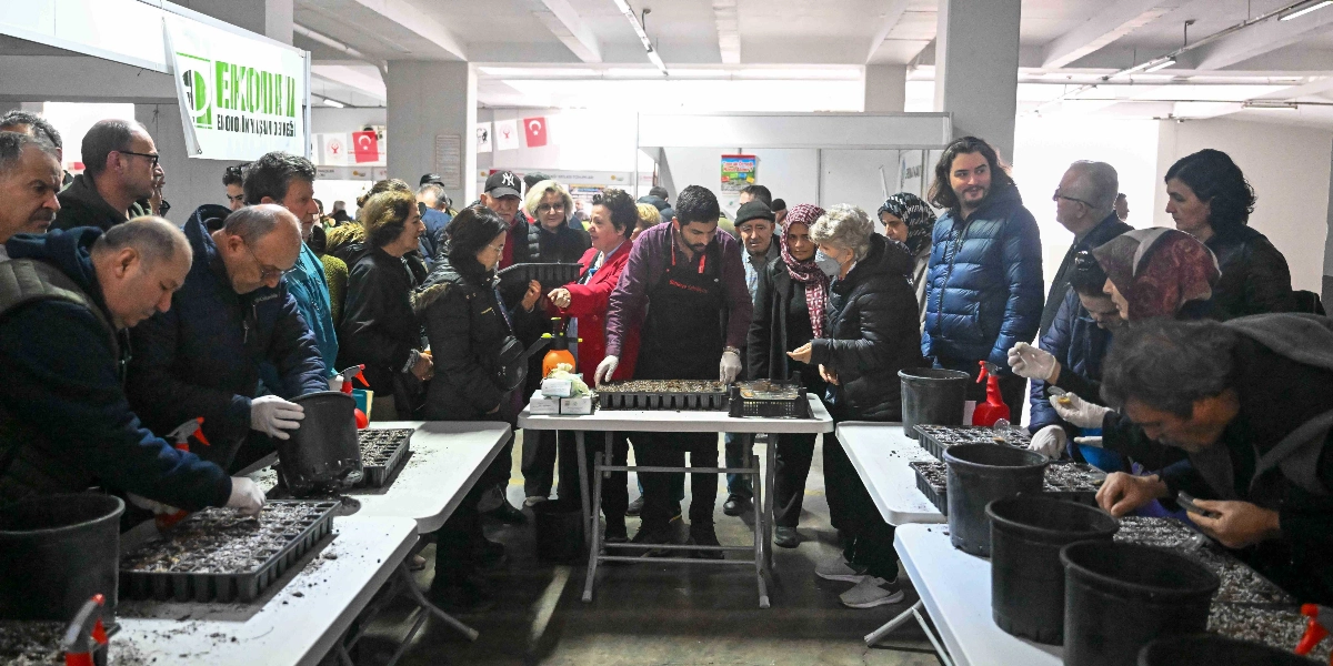Kapalı bir etkinlik alanında, beyaz masaların etrafında toplanmış bir grup insan, yerel tohum ekimi eğitimi alıyor. Masaların üzerinde toprak dolu viyoller, saksılar ve sprey şişeleri bulunuyor. Merkezde duran bir eğitmen, katılımcılara tohum ekimini uygulamalı olarak gösterirken, çevresindeki farklı yaş gruplarından katılımcılar dikkatle izliyor ve bazıları kendi viyollerine tohum ekiyor. Arka planda üzerinde 
