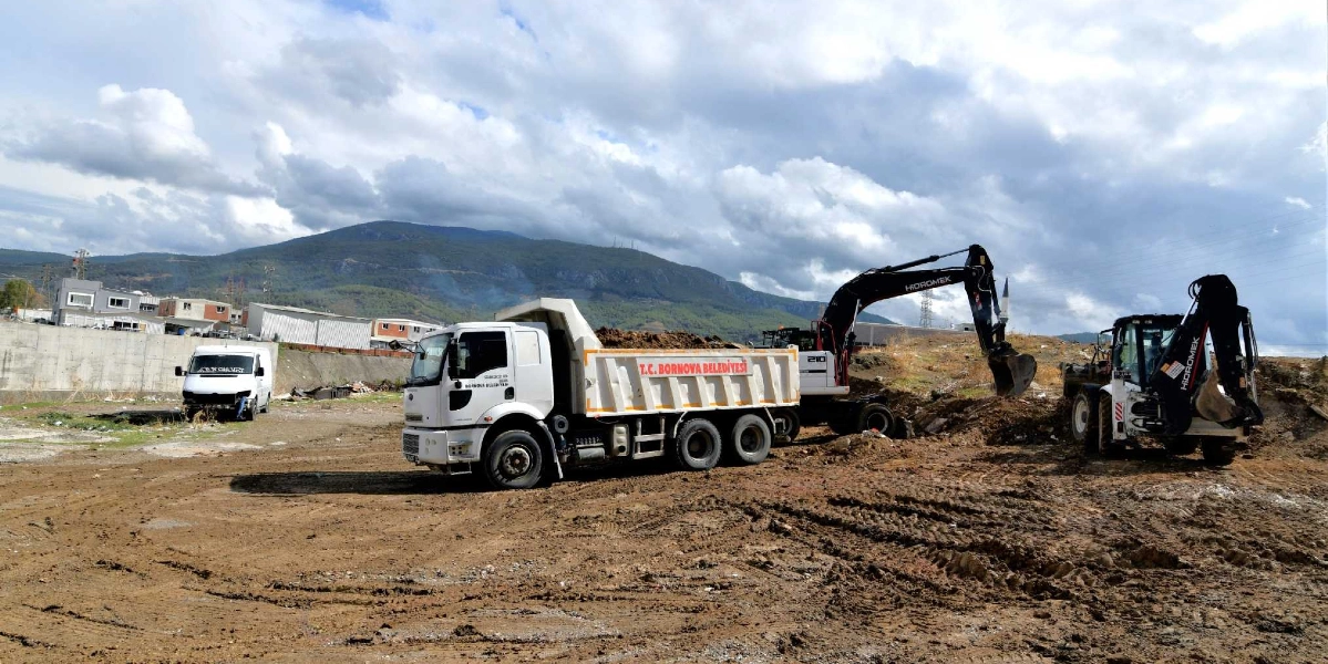 T.C. Bornova Belediyesi'ne ait bir damperli kamyon, bir ekskavatör ve bir kepçenin, dağ manzaralı geniş ve çamurlu bir arazide toprak düzenleme çalışması yaptığı görülüyor.