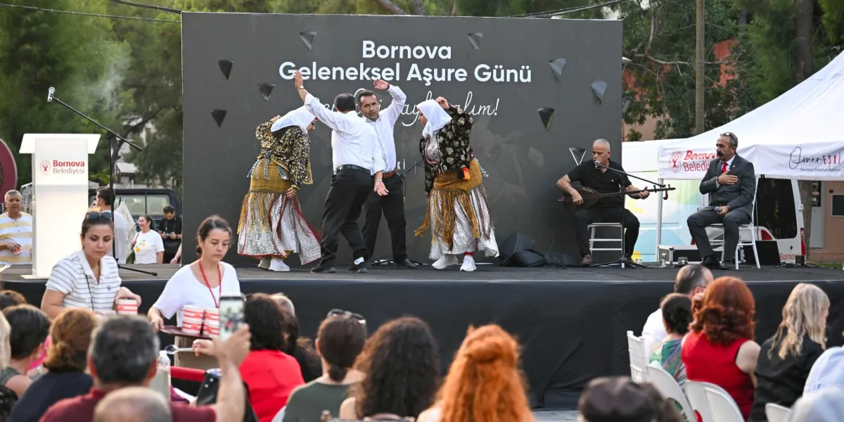 Bornova Belediyesi tarafından düzenlenen Geleneksel Aşure Günü etkinliğinde, sahne üzerinde geleneksel kıyafetli iki kadın ve iki erkek halk dansı gösterisi sergiliyor. Sahnenin sağ tarafında bağlama çalan bir müzisyen ve yanında elini kalbine koymuş oturan Bornova Belediye Başkanı Ömer Eşki yer alıyor. Sahne arkasındaki panoda 