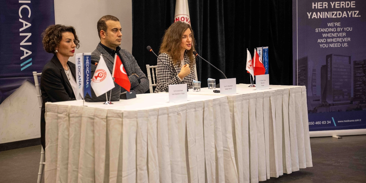 Beyaz örtülü uzun bir masada yan yana oturan üç konuşmacı, rahim ağzı kanseri farkındalık etkinliği kapsamında bir panelde yer alıyor. En sağda oturan kadın konuşmacı mikrofon başında açıklamalarda bulunurken, yanında bir erkek ve bir başka kadın konuşmacı onu dinliyor. Masanın üzerinde küçük Türk bayrakları, kurum logolu flamalar ve konuşmacıların isimlerinin yer aldığı kartlar bulunuyor. Arka planda ise üzerinde 