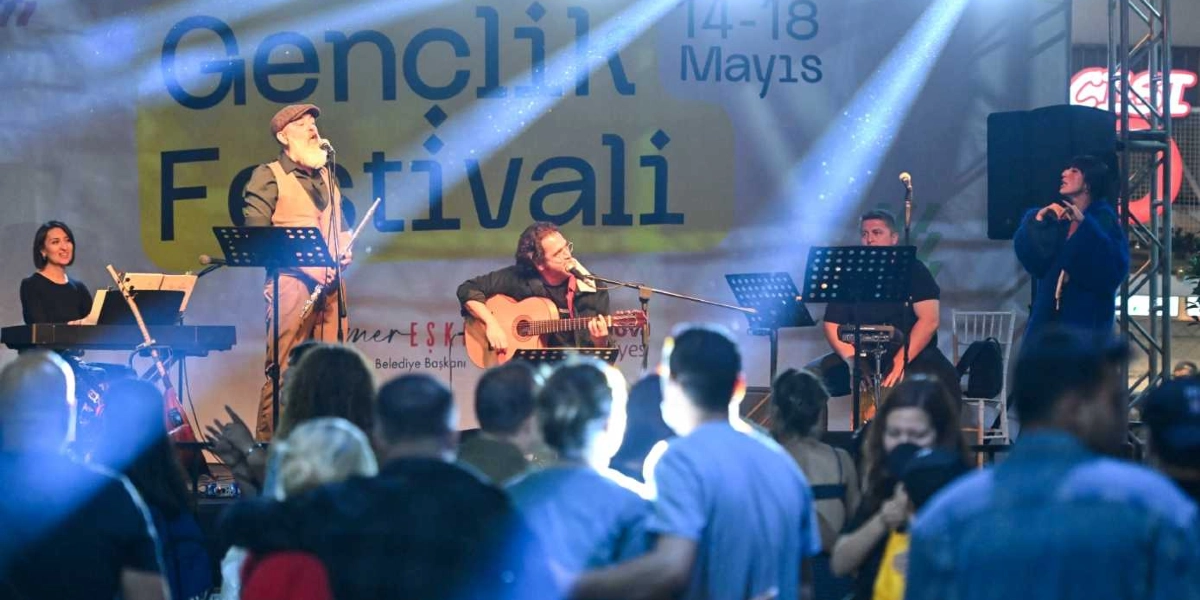 Küçükpark'ta düzenlenen Gençlik Festivali sahnesinde, arkada üzerinde 14-18 Mayıs tarihleri yazılı büyük bir afişin önünde müzik grubu performans sergiliyor. Sahnede piyano başında bir kadın, ayakta şarkı söyleyen şapkalı bir erkek, gitar çalan bir erkek, perküsyon başında oturan bir erkek ve mikrofonla şarkı söyleyen mavi ceketli bir kadın yer alıyor. Sahne ışıklarıyla aydınlatılan grubun önünde, konseri izleyen kalabalık bir izleyici kitlesi bulunuyor.