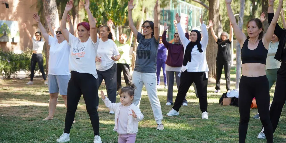 Açık havada, yeşil bir alanda spor kıyafetleri giymiş bir grup kadın, ellerini havaya kaldırarak egzersiz yapıyor. Grubun ön tarafında, diğerlerine eşlik eder gibi ellerini iki yana açmış küçük bir kız çocuğu bulunuyor. Katılımcıların çoğu spor tişörtleri ve taytlar giymiş, neşeli bir atmosferde hareket ediyor.