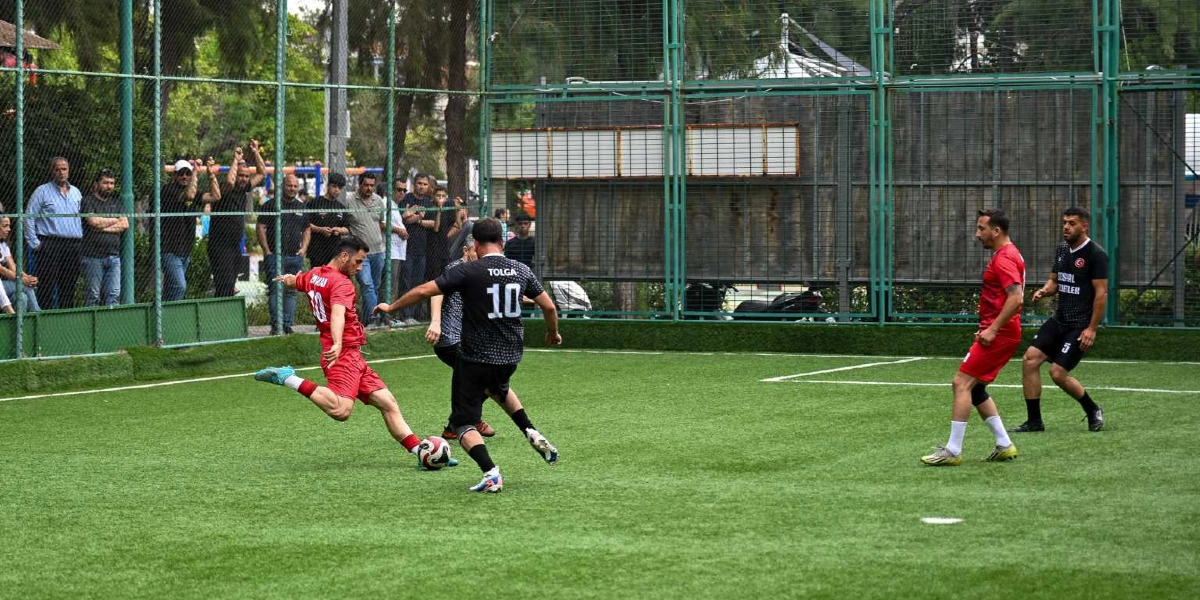 Açık havada, tel örgülerle çevrili suni çim sahada futbol oynayan dört oyuncu. Kırmızı formasıyla topa vuran bir oyuncu, onu karşılayan siyah formasıyla sırtında 10 numara yazan Tolga, sağ tarafta bekleyen kırmızı formalı başka bir oyuncu ve siyah formasıyla izleyen bir diğer oyuncu sahada yer alıyor. Arka planda tel örgülerin arkasında maçı izleyen bir grup seyirci bulunuyor.