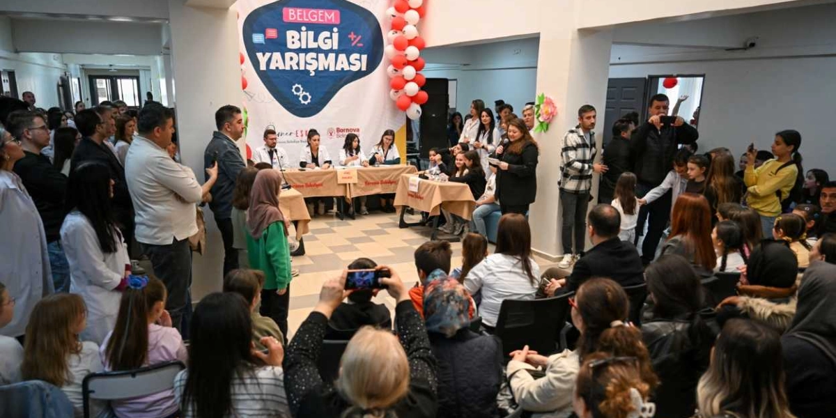 Bornova Belediyesi tarafından düzenlenen BELGEM Bilgi Yarışması etkinliğinde, salonu dolduran kalabalık bir izleyici grubu ve ön planda yarışmacıların oturduğu masalar görülüyor. Arka planda üzerinde 
