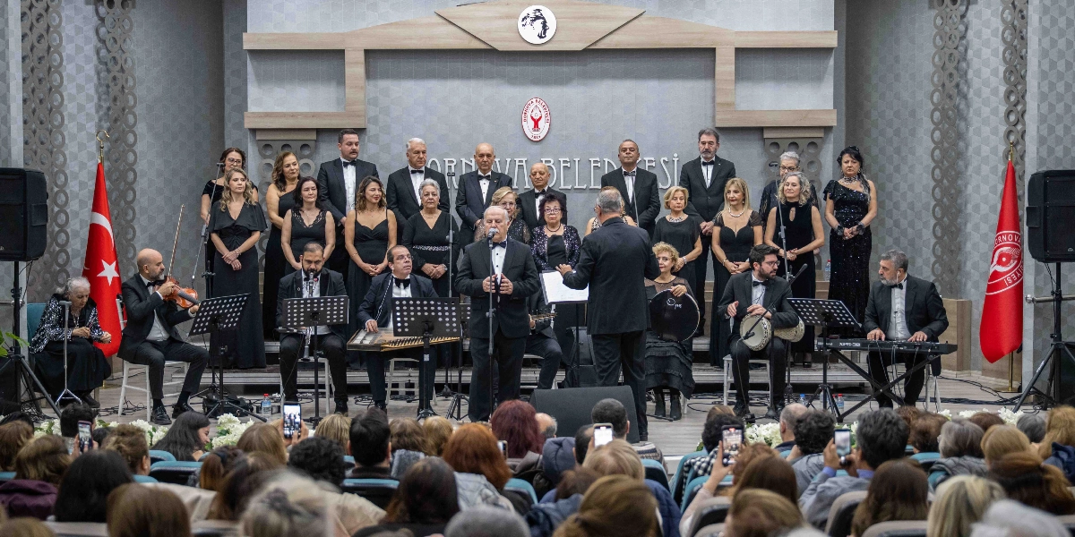 Bornova Belediyesi'ne ait bir sahnede, siyah kıyafetler giymiş bir koro ve müzisyen grubu performans sergiliyor. Arka planda Bornova Belediyesi logosu ve Atatürk silüeti yer alırken, yanlarda Türk bayrağı ve belediye flaması bulunuyor. Ön planda şefin yönetiminde şarkı söyleyen solist, kanun, keman, darbuka ve klavye çalan müzisyenler ile koro üyeleri görülüyor. İzleyiciler ise sahneyi karşıdan izliyor ve bazıları telefonlarıyla kayıt yapıyor.