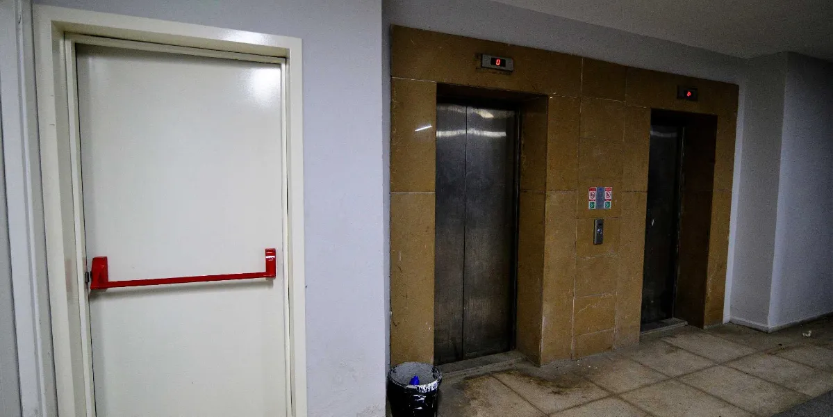 Gri duvarlı bir koridorda, sol tarafta kırmızı panik barı olan beyaz bir yangın çıkış kapısı, sağ tarafta ise sarı mermer kaplamalı iki adet metal asansör kabini bulunuyor. Asansörlerin üzerinde dijital kat göstergeleri yer alıyor ve asansörlerin önünde siyah bir çöp kovası duruyor.