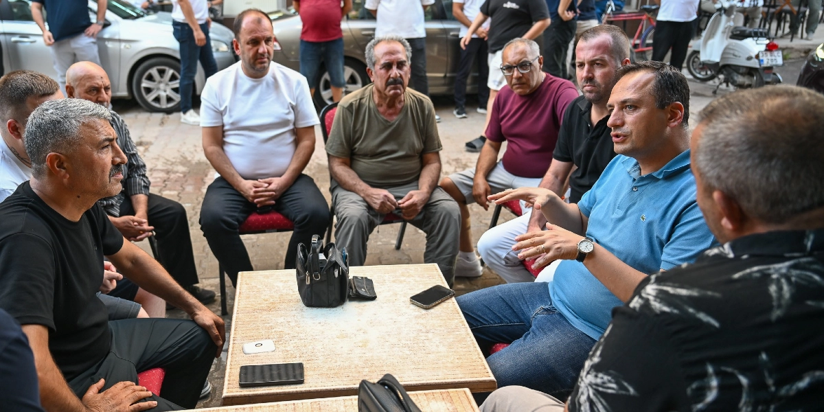 Belediye Başkanı, açık havada bir masanın etrafında toplanmış esnafla sohbet ediyor. Mavi polo yaka tişört giymiş olan Başkan, el hareketleriyle bir şeyler anlatırken, çevresindeki erkeklerden oluşan grup onu dikkatle dinliyor. Masanın üzerinde birkaç cep telefonu ve küçük bir çanta bulunuyor. Arka planda park halindeki araçlar ve sokaktaki diğer insanlar görülüyor.
