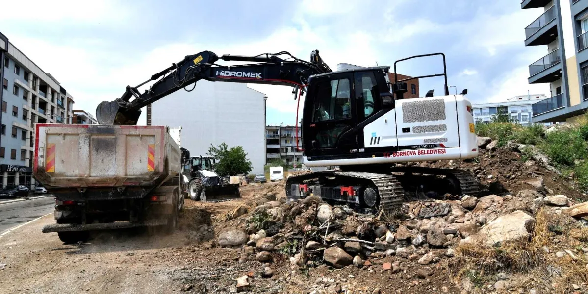 Üzerinde T.C. Bornova Belediyesi yazılı beyaz ve siyah renkli Hidromek marka paletli ekskavatör, bir inşaat alanında toprak ve moloz yığınlarını temizleyerek arkasındaki damperli kamyona yüklüyor.