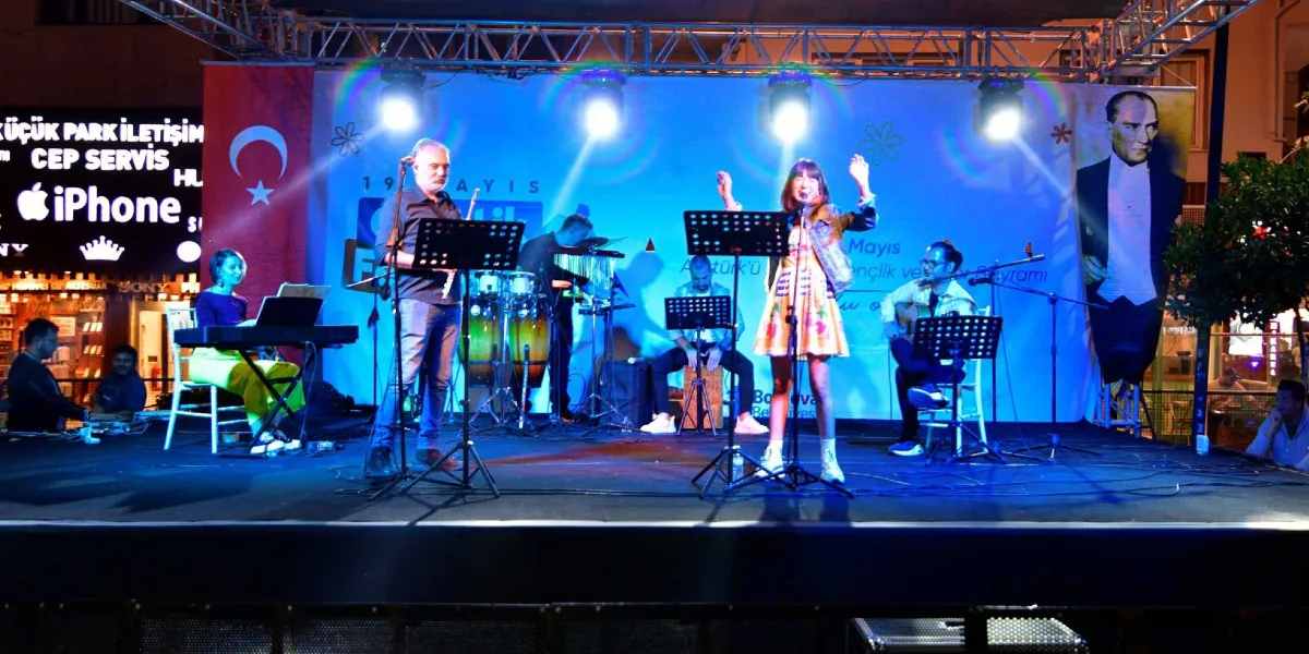 Gece vakti açık havada kurulan bir sahnede müzik grubu konser veriyor. Sahnenin arka planında üzerinde Türk bayrağı, Atatürk portresi ve 19 Mayıs Gençlik ve Spor Bayramı yazılı afişler bulunuyor. Mavi ışıklarla aydınlatılan sahnede, ayakta şarkı söyleyen kadın solist, klavye çalan bir kadın, gitarist, perküsyoncu ve diğer müzisyenler yer alıyor. Müzisyenlerin önünde nota sehpaları bulunuyor ve sahne profesyonel ışıklandırma sistemleriyle aydınlatılıyor.