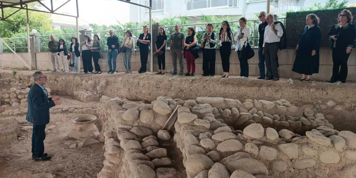 Açık hava arkeolojik kazı alanında, takım elbiseli bir konuşmacı, kazı alanının kenarında sıralanmış yaklaşık yirmi kişilik bir gruba bilgi veriyor. Arkeolojik kalıntıların arasında büyük taş bloklar ve toprak yapılar dikkat çekerken, katılımcılar merakla konuşmacıyı dinliyor.