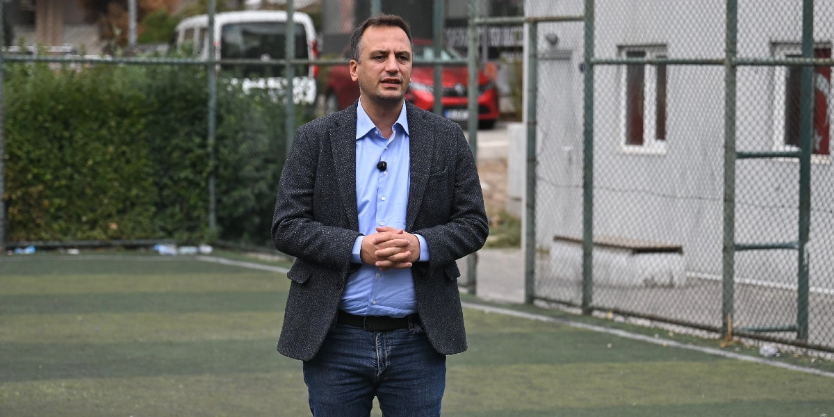 Mavi gömlek, koyu renk ceket ve kot pantolon giymiş bir erkek, açık hava spor sahasında ellerini önünde birleştirmiş şekilde konuşma yapıyor. Arka planda tel örgülerle çevrili bir saha ve park halindeki kırmızı bir otomobil görülüyor.