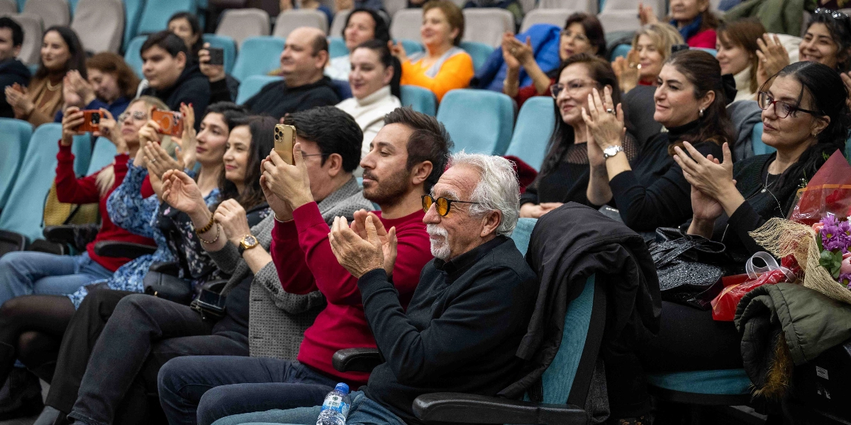 Bir konser veya etkinlik salonunda, mavi koltuklarda oturan izleyiciler alkışlayarak sahneyi izliyor. Ön planda, kırmızı kazaklı bir adam cep telefonuyla video kaydederken, yanında beyaz saçlı, gözlüklü ve siyah kıyafetli bir adam alkışlıyor. İzleyicilerin bir kısmı telefonlarıyla çekim yaparken, bir kısmı da ellerinde çiçeklerle sahnedeki performansı keyifle takip ediyor.