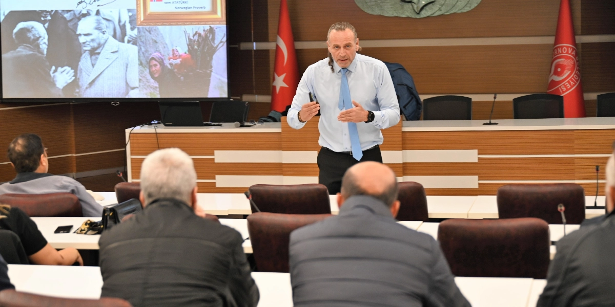 Resmi bir toplantı salonunda, açık mavi gömlekli ve açık mavi kravatlı bir konuşmacı, dinleyicilere hitap ederek sunum yapıyor. Konuşmacının arkasındaki ekranda Atatürk'ün bir fotoğrafı ve sırtında odun taşıyan bir kadın görseli yer alıyor. Salonun arka planında Türk bayrağı ve Bornova Belediyesi'nin logosunun bulunduğu bir bayrak bulunuyor; ön planda ise sırtları dönük şekilde oturan dinleyiciler görünüyor.