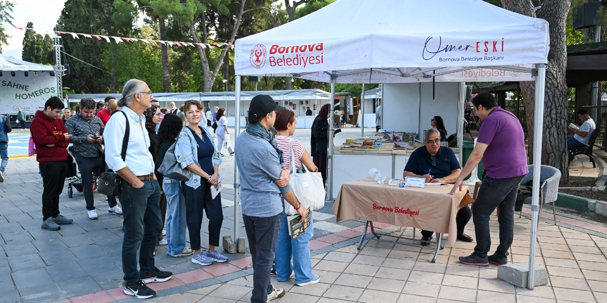 Bornova Belediyesi tarafından düzenlenen bir etkinlikte, üzerinde Bornova Belediyesi ve Bornova Belediye Başkanı Ömer Eşki'nin logolarının bulunduğu beyaz bir çadırın önünde insanlar kitap imzalatmak için sıra bekliyor. Çadırın içindeki masada oturan yazar, okurları için kitap imzalarken, mor tişörtlü bir kişi de masanın yanında duruyor. Etkinlik alanının arka planında ağaçlar ve başka stantlar görülüyor.