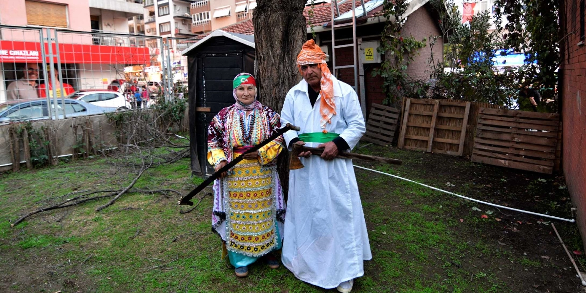 Geleneksel kıyafetler giymiş bir kadın ve bir erkek, bir ağacın önünde yan yana duruyor. Kadın, üzerinde renkli işlemeler ve pullar bulunan yöresel bir elbise, başında Türk bayrağı motifli bir başörtüsü ile elinde uzun bir testere tutuyor. Yanındaki erkek, beyaz uzun bir entari, belinde yeşil bir kuşak ve başında turuncu bir sarıkla elinde bir balta taşıyor. Arka planda bir bahçe, ahşap paletler ve şehir merkezini andıran binalar görülüyor.