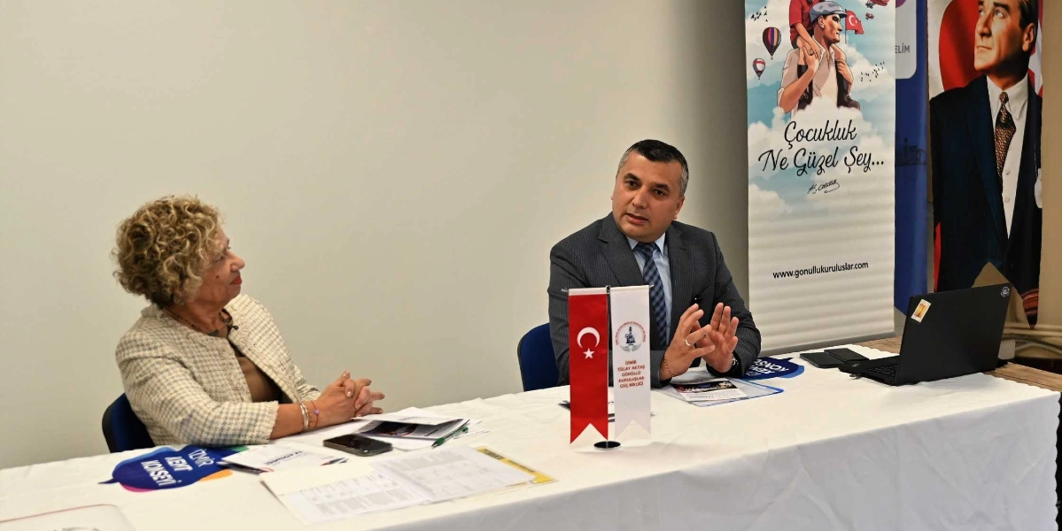Beyaz bir masa etrafında oturan iki kişi, bir toplantı sırasında konuşuyor. Masanın sağ tarafında gri takım elbiseli ve kravatlı bir erkek, ellerini kullanarak bir şeyler anlatıyor. Masanın sol tarafında ise kıvırcık sarı saçlı, bej renkli ceket giyen bir kadın, konuşmacıyı dinliyor. Masanın üzerinde Türk bayrağı ve İzmir Tülay Aktaş Gönüllü Kuruluşlar Güç Birliği yazılı küçük bir masa bayrağı, bazı belgeler ve açık bir dizüstü bilgisayar bulunuyor. Arka planda ise üzerinde 