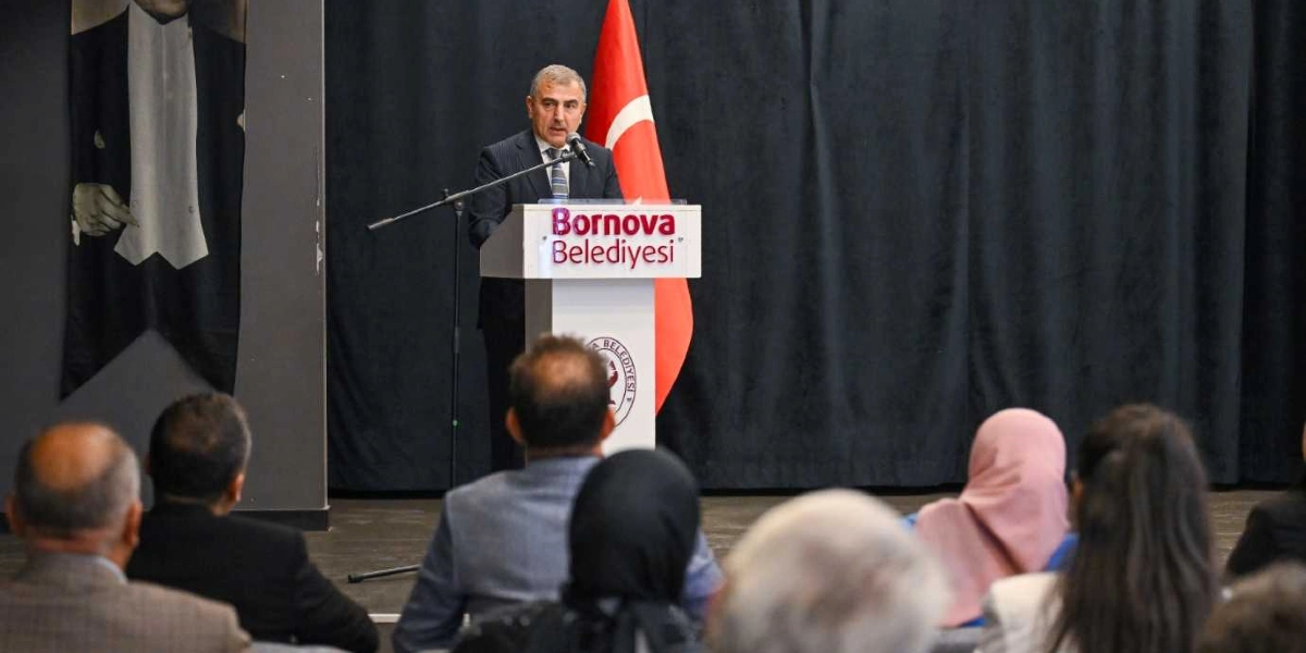 Koyu renkli bir perde önünde, üzerinde Bornova Belediyesi logosu ve yazısı bulunan bir kürsüde konuşma yapan takım elbiseli bir erkek, karşısında oturan dinleyici grubuna hitap ediyor. Kürsünün hemen arkasında Türk bayrağı, sol tarafta ise Atatürk portresinin bir kısmı görülüyor.