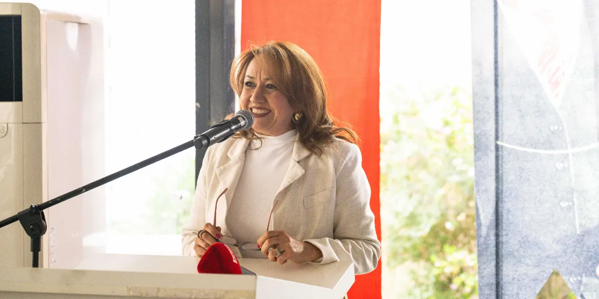 Krem rengi ceket ve beyaz boğazlı bir kazak giymiş, omuz hizasında kahverengi saçlı bir kadın, kürsüde mikrofon önünde konuşma yapıyor. Kadın, elinde gözlük tutuyor ve gülümseyerek ileriye bakıyor. Arka planda kırmızı bir panel ve aydınlık bir iç mekan bulunuyor.