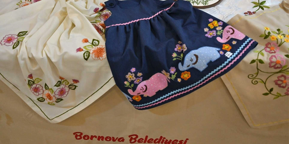 Krem rengi kumaşlar üzerine işlenmiş çiçek motifleri ve ortada üzerinde fil figürleri ile çiçek işlemeleri bulunan lacivert bir çocuk elbisesi yer alıyor. Alt kısımda kırmızı harflerle Bornova Belediyesi yazısı görülüyor.
