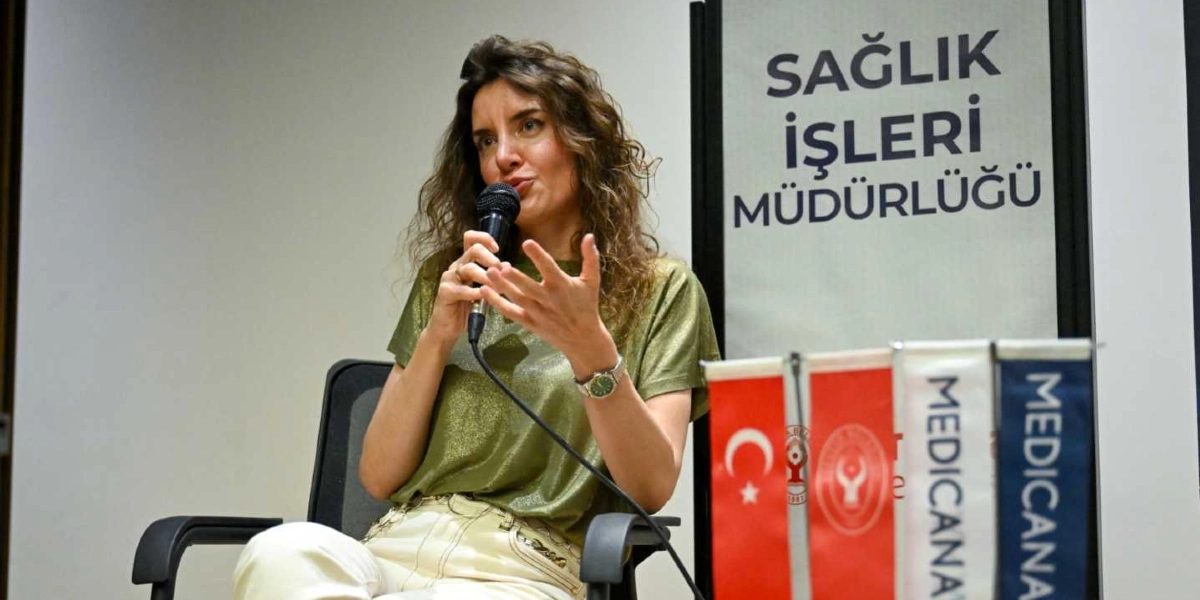 Kıvırcık saçlı bir kadın, elinde mikrofonla konuşma yapıyor. Kadın, parlak yeşil bir bluz ve açık renkli pantolon giymiş, bir elini jest yaparak havaya kaldırmış durumda. Arka planda üzerinde 