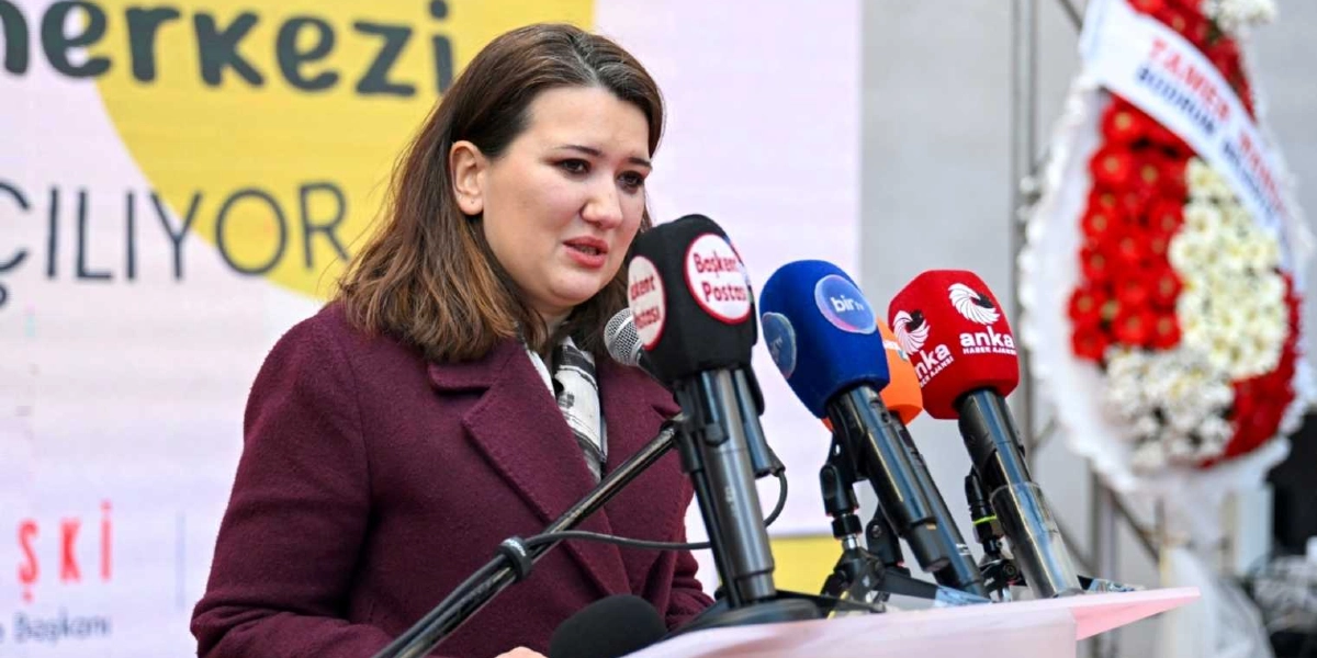 Bordo renkli kaban giymiş bir kadın, önünde dizili olan Başkent Postası, Bir TV ve Anka Haber Ajansı mikrofonlarına konuşma yapıyor. Arka planda üzerinde 