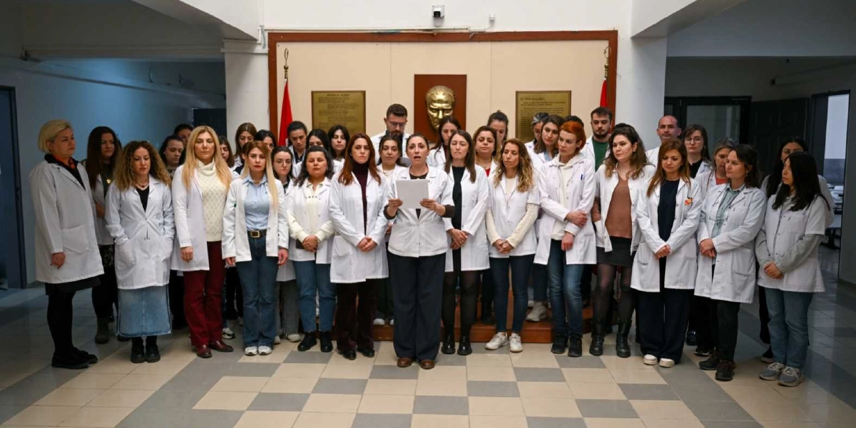 Beyaz önlük giymiş kalabalık bir öğretmen grubu, Bornova Belediyesi Gençlik Eğitim Merkezi'nde (BELGEM) şiddeti protesto etmek amacıyla bir araya gelmiş. Grubun ortasında duran bir kadın, elindeki yazılı metni okuyarak basın açıklaması yapıyor. Arka planda duvarda Atatürk'ün kabartma portresi ve iki yanında Türk bayrakları yer alıyor.
