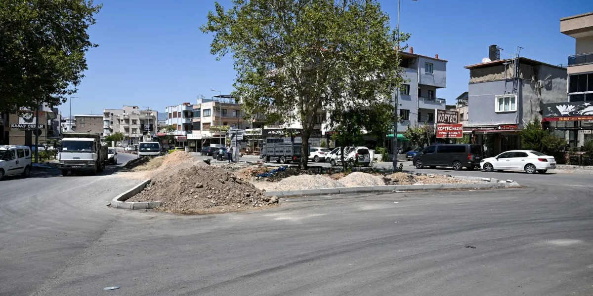 Pınar Caddesi'nde devam eden yol yenileme çalışmaları kapsamında, caddenin ortasındaki refüj alanında toprak ve moloz yığınları bulunuyor. Çevredeki binaların önünde park etmiş araçlar, iş makineleri ve cadde boyunca sıralanan dükkanlar görülüyor. Gökyüzü açık ve güneşli bir gün.