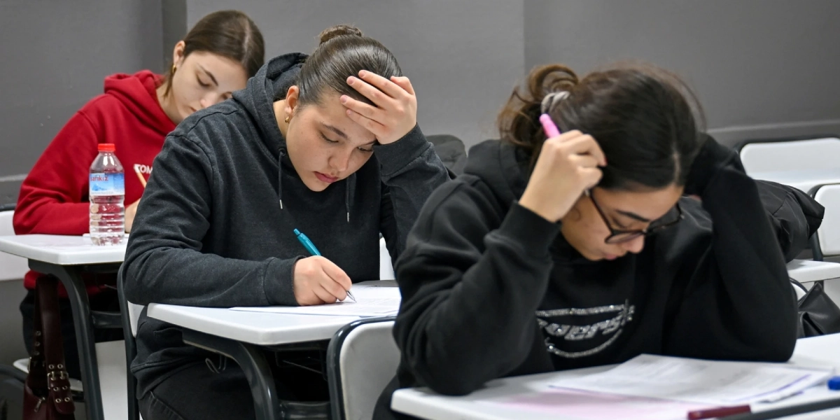 Sınıf ortamında sınav olan üç genç kız, sıralarında oturmuş dikkatle önlerindeki kağıtlara odaklanmış durumdalar. En öndeki gözlüklü öğrenci elinde pembe bir kalemle düşünceli bir şekilde kağıdına bakarken, yanındaki öğrenci elini alnına koymuş yazı yazıyor. Arka planda kırmızı sweatshirt giyen bir diğer öğrenci ise sınav kağıdına odaklanmış şekilde çalışıyor.