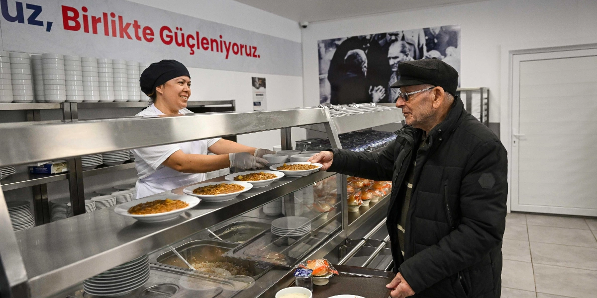 Bir yemekhanede, beyaz önlüklü ve şapkalı bir kadın görevli, tezgahın arkasından yaşlı bir adama içinde yemek olan bir tabağı uzatıyor. Yaşlı adam, elinde tepsisiyle yemeği teslim alırken gülümsüyor. Arka planda duvarda 
