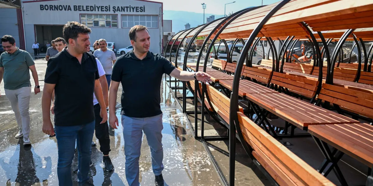 Bornova Belediyesi Şantiyesi önünde, siyah tişörtlü bir yetkili, parklar için tasarlanmış, siyah metal iskeletli ve ahşap oturma bölümlerinden oluşan tünel formundaki yeni nesil oturma gruplarını yanındaki kişilere tanıtıyor.