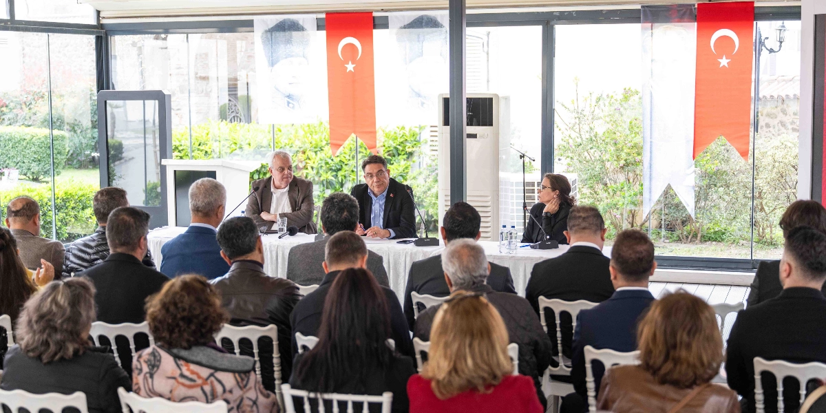 CHP İzmir Milletvekili Yankı Bağcıoğlu'nun Bornova'da gerçekleştirdiği toplantıda, beyaz örtülü bir masada konuşma yaptığı ve katılımcıların kendisini dinlediği bir etkinlikten görünüm. Arka planda cam bölmelerin önünde asılı Türk bayrakları ve Atatürk posterleri yer alıyor.