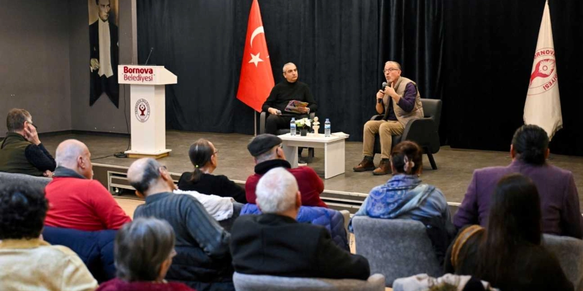Bornova Belediyesi'nin düzenlediği bir etkinlikte, sahnede iki konuşmacı izleyicilere hitap ediyor. Sahnede, üzerinde Bornova Belediyesi logosu bulunan bir kürsü, Türk bayrağı ve Bornova Belediyesi'nin flaması yer alıyor. Konuşmacılardan biri elinde mikrofonla ayakta dururken diğeri yanında oturuyor. Salonu dolduran izleyiciler, sahnedeki konuşmacıları dikkatle dinliyor. Arka planda duvarda Mustafa Kemal Atatürk'ün bir portresi asılı duruyor.