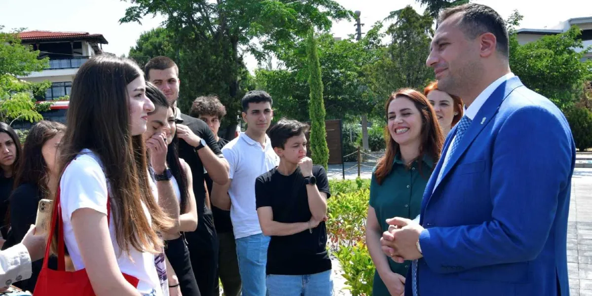 Mavi takım elbiseli bir erkek, açık havada bir grup gençle sohbet ediyor. Gençler ilgiyle dinlerken, grubun yanındaki bir kadın gülümseyerek onları izliyor. Arka planda ağaçlar ve bir bina yapısı görülüyor.