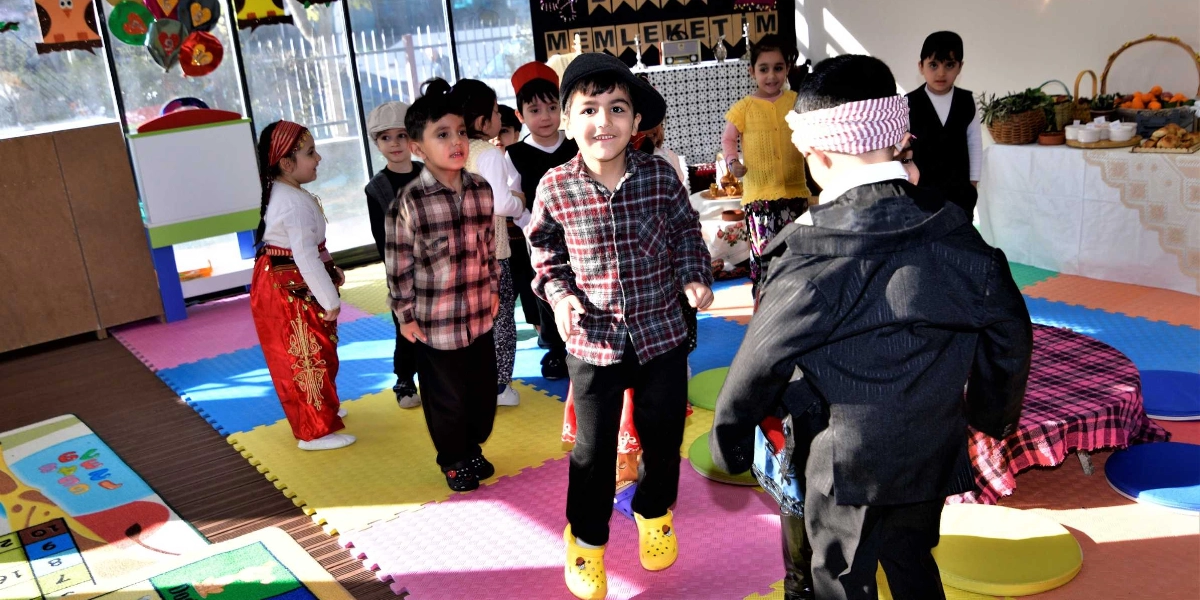 Yerli Malı Haftası kutlaması kapsamında geleneksel kıyafetler giymiş anaokulu öğrencileri, renkli yer minderleri ve oyun matlarıyla kaplı bir sınıfta bir arada bulunuyor. Ön planda ekose gömlekli ve şapkalı bir erkek çocuk gülümseyerek kameraya bakarken, çevresinde yöresel kıyafetli kız ve erkek çocuklar yer alıyor. Arka planda üzerinde 