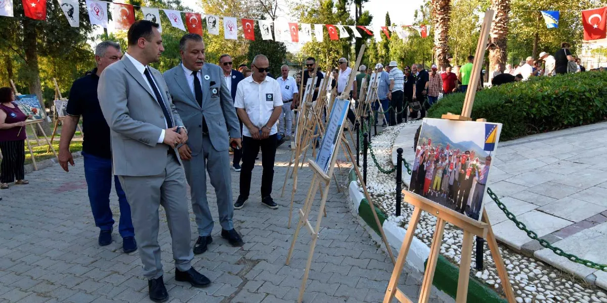 Açık havada, park benzeri bir alanda düzenlenen Srebrenica anma sergisinde, takım elbiseli yetkililer ve katılımcılar şövale üzerindeki fotoğrafları inceliyor. Arka planda Türk bayraklarıyla süslenmiş bir yürüyüş yolu ve ağaçlar yer alırken, ön planda yer alan bir şövale üzerinde Srebrenica'yı anımsatan bir grup fotoğrafı sergileniyor.