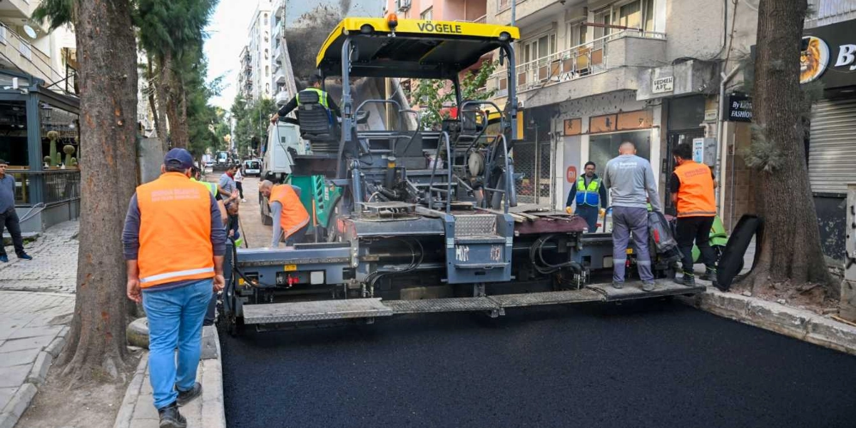 Süvari Caddesi'nde asfalt serme çalışması yapan işçiler ve büyük bir finişer makinesi. Turuncu yelekli belediye çalışanları yolun kenarında ve makinenin üzerinde görev yaparken, arkada bir kamyon ve çevrede apartmanlar ile dükkanlar görülüyor.