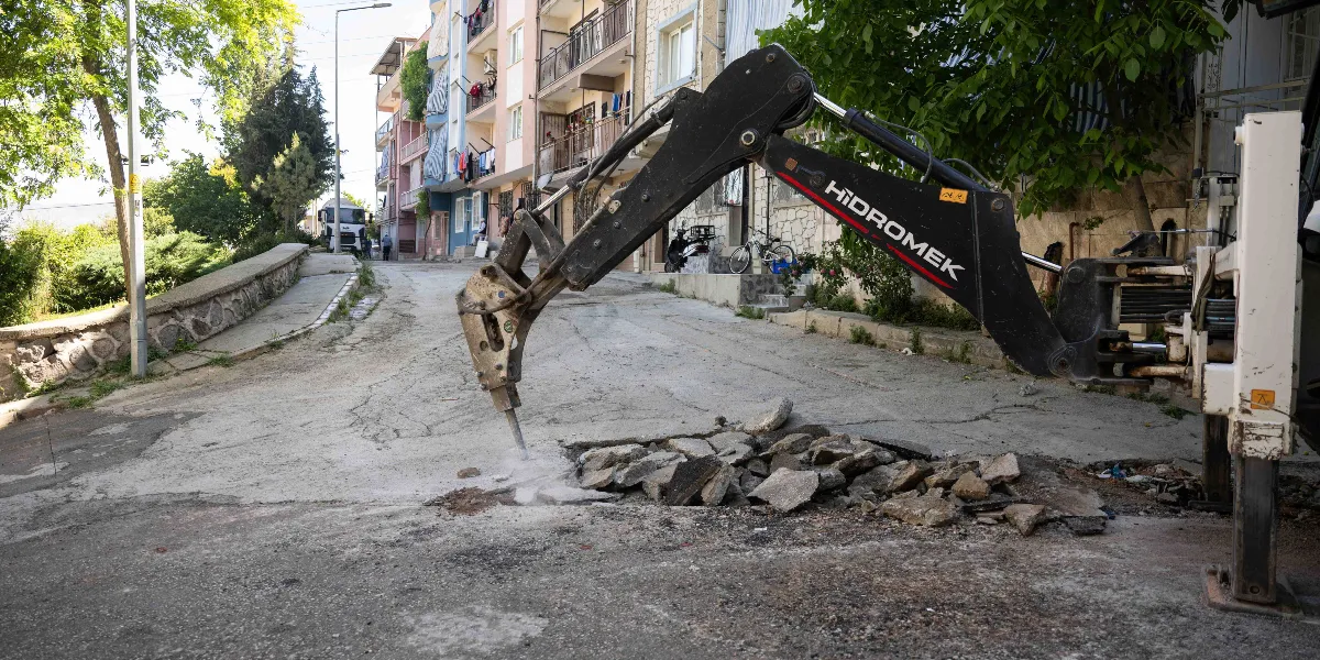 Bornova'da eğimli bir sokakta, Hidromek marka bir iş makinesi asfalt zemini kırmak için çalışıyor. Kırıcı uç, yol üzerindeki eski asfaltı parçalarken etrafa toz yayılıyor. Arka planda apartmanlar, ağaçlar ve park halindeki bir bisiklet görülüyor.