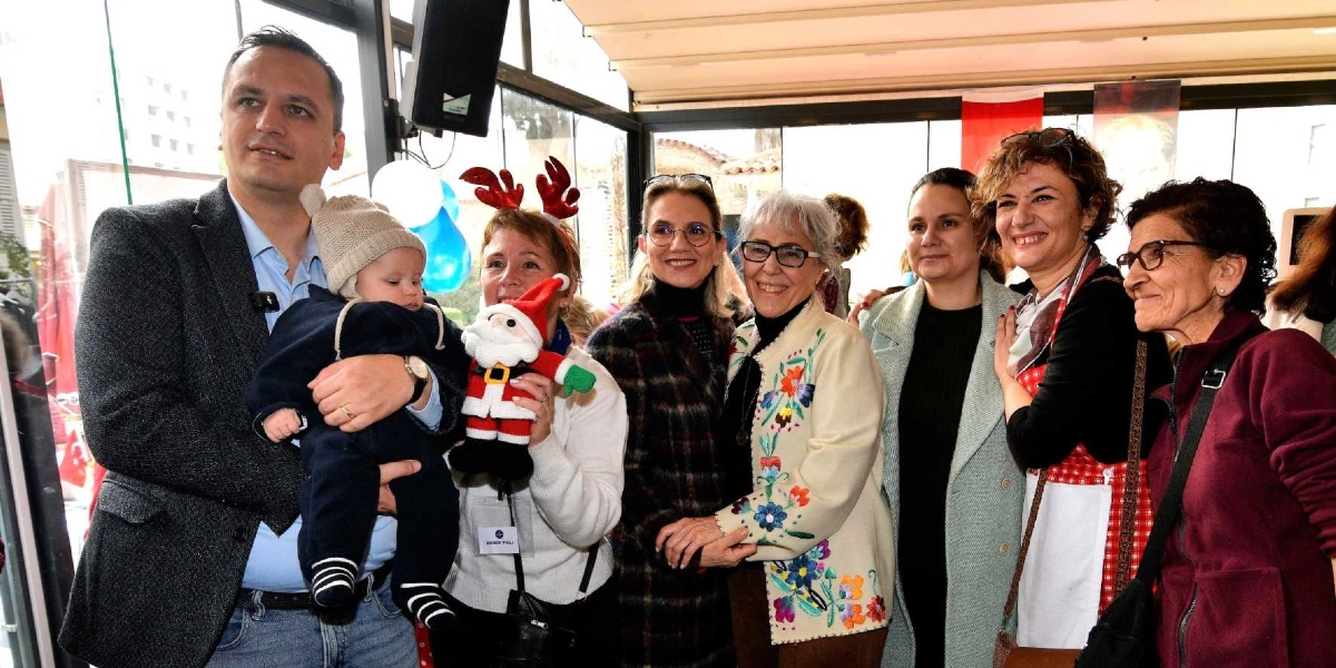 Kapalı bir mekanda bir araya gelen bir grup insan, gülümseyerek kameraya bakıyor. Sol tarafta bir erkek, kucağında bebekle duruyor. Yanındaki kadın, başında geyik boynuzlu bir taç takmış ve elinde Noel Baba figürü tutuyor. Diğer kadınlar ise yan yana dizilmiş, samimi bir şekilde poz veriyor. İçlerinden biri çiçek işlemeli bir ceket, bir diğeri ise üzerinde kırmızı beyaz ekose desenli bir önlük giyiyor. Arka planda cam bölmelerin ardında dış mekan ve kısmen görünen bayraklar yer alıyor.