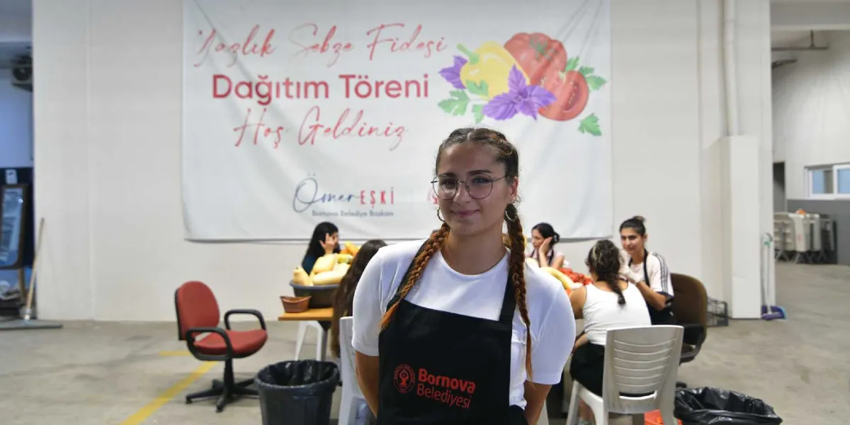 Beyaz tişört ve üzerinde Bornova Belediyesi logosu bulunan siyah bir önlük giymiş, saçları örgülü genç bir kadın kameraya gülümseyerek bakıyor. Arka planda 