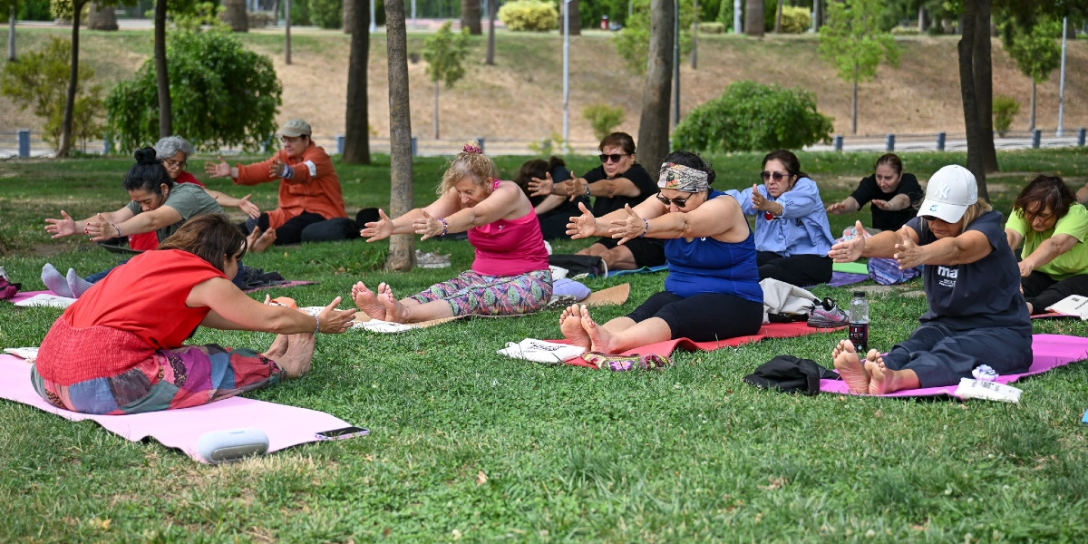 Ağaçlıklı bir parkta, çimlerin üzerine serilmiş matlar üzerinde yoga yapan bir grup kadın, bacaklarını öne uzatıp kollarını ileriye doğru esneterek oturma pozunda egzersiz yapıyor. Farklı renklerde spor kıyafetleri giymiş kadınlar, açık havada bir eğitmen eşliğinde grup halinde spor yapıyor.