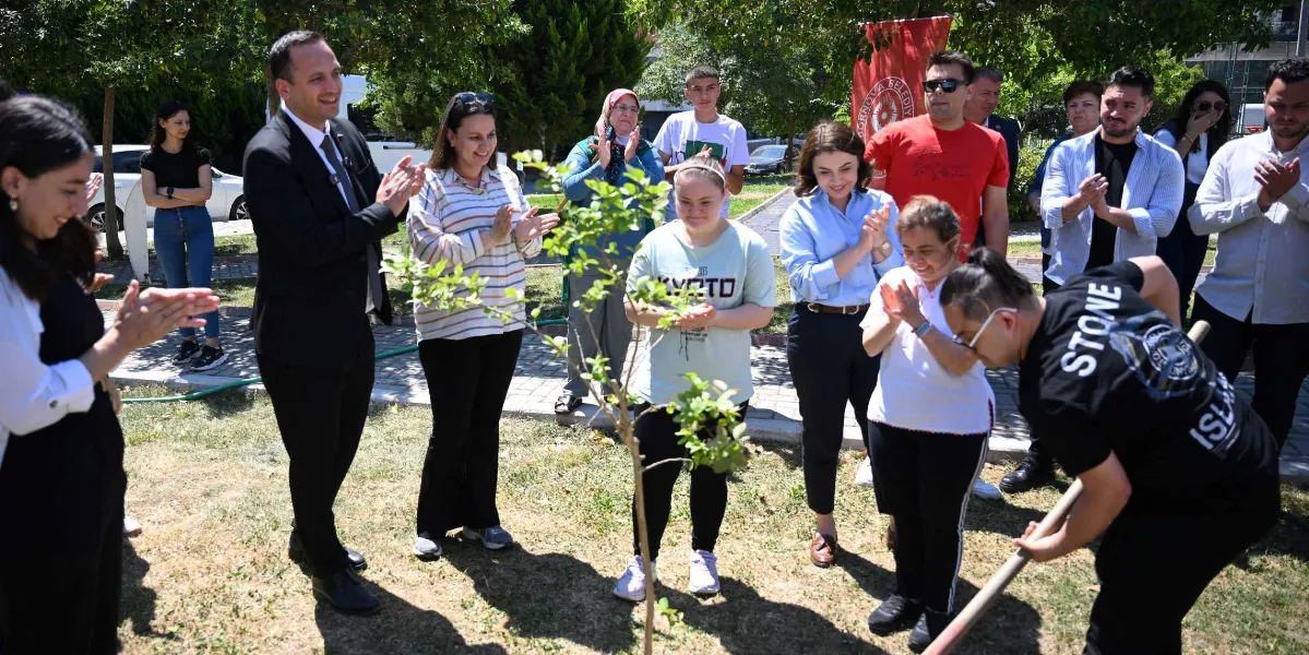 Açık havada, yeşil bir alanda düzenlenen fidan dikme etkinliğinde, siyah tişörtlü bir genç elindeki kürekle yeni dikilen bir fidanın toprağını düzeltiyor. Etrafında toplanan, aralarında Bornova Belediyesi yetkililerinin de bulunduğu kalabalık bir grup, genci alkışlayarak destek veriyor. Katılımcılar arasında down sendromlu bireylerin de yer aldığı grup, neşeli bir şekilde fidan dikimini izliyor. Arka planda Bornova Belediyesi logolu kırmızı bir bayrak ve ağaçlar görülüyor.