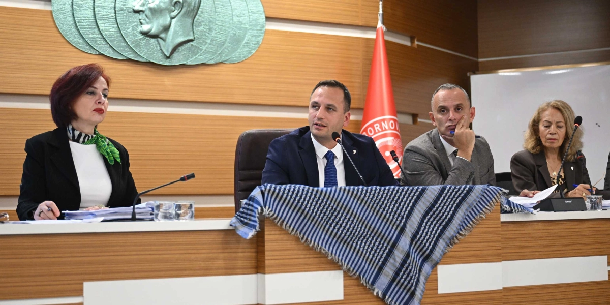 Bornova Belediye Meclisi toplantısında, kürsüde oturan dört kişi Filistin'e destek amacıyla masanın önüne serilmiş mavi beyaz çizgili bir Filistin kefiyesi ile oturuyor. Arka planda Bornova Belediyesi'nin bayrağı ve duvarda Atatürk silüeti bulunan bir kabartma yer alıyor.