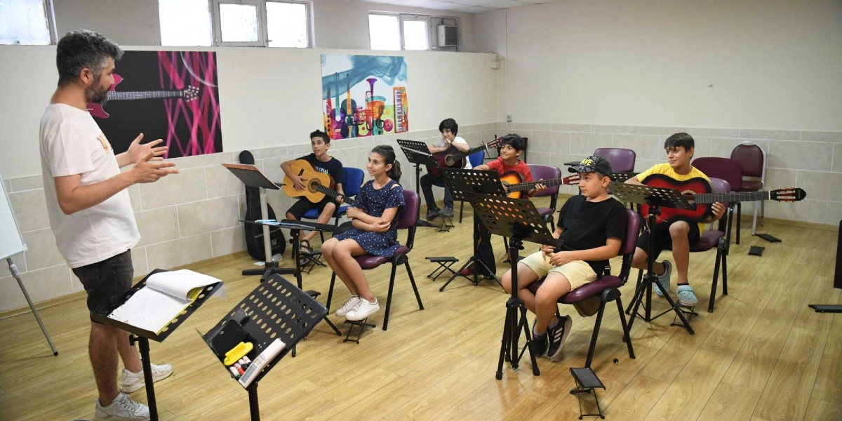 Sınıf ortamında gitar eğitimi veren sakallı bir eğitmen ve önlerinde nota sehpalarıyla oturarak gitar çalan beş çocuk. Eğitmen ayakta öğrencilere bir şeyler anlatırken, öğrenciler sandalyelerinde oturmuş, kucaklarında gitarlarıyla derse odaklanmış durumdalar. Duvarlarda müzik temalı renkli tablolar asılı ve zemin ahşap parke ile kaplı.